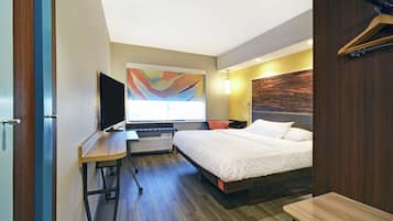 Habitación de hotel con cama, escritorio con televisor de pantalla plana, calefactor, ventana con un cuadro abstracto de colores y cabecera de madera.