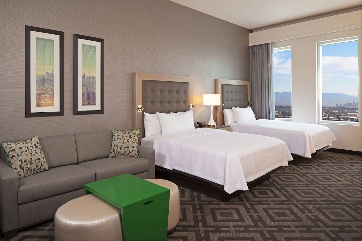 Ein Hotelzimmer mit zwei Betten, einem Schlafsofa, einem grünen Tisch und Blick auf die Stadt.