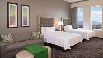 Quarto de hotel com duas camas, um sofá, uma mesa verde e vista para a cidade.