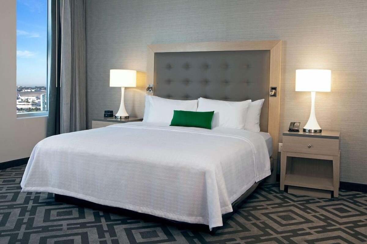 A hotel room with a large bed, two bedside tables with lamps, a green accent pillow, and a view of the city through a large window.