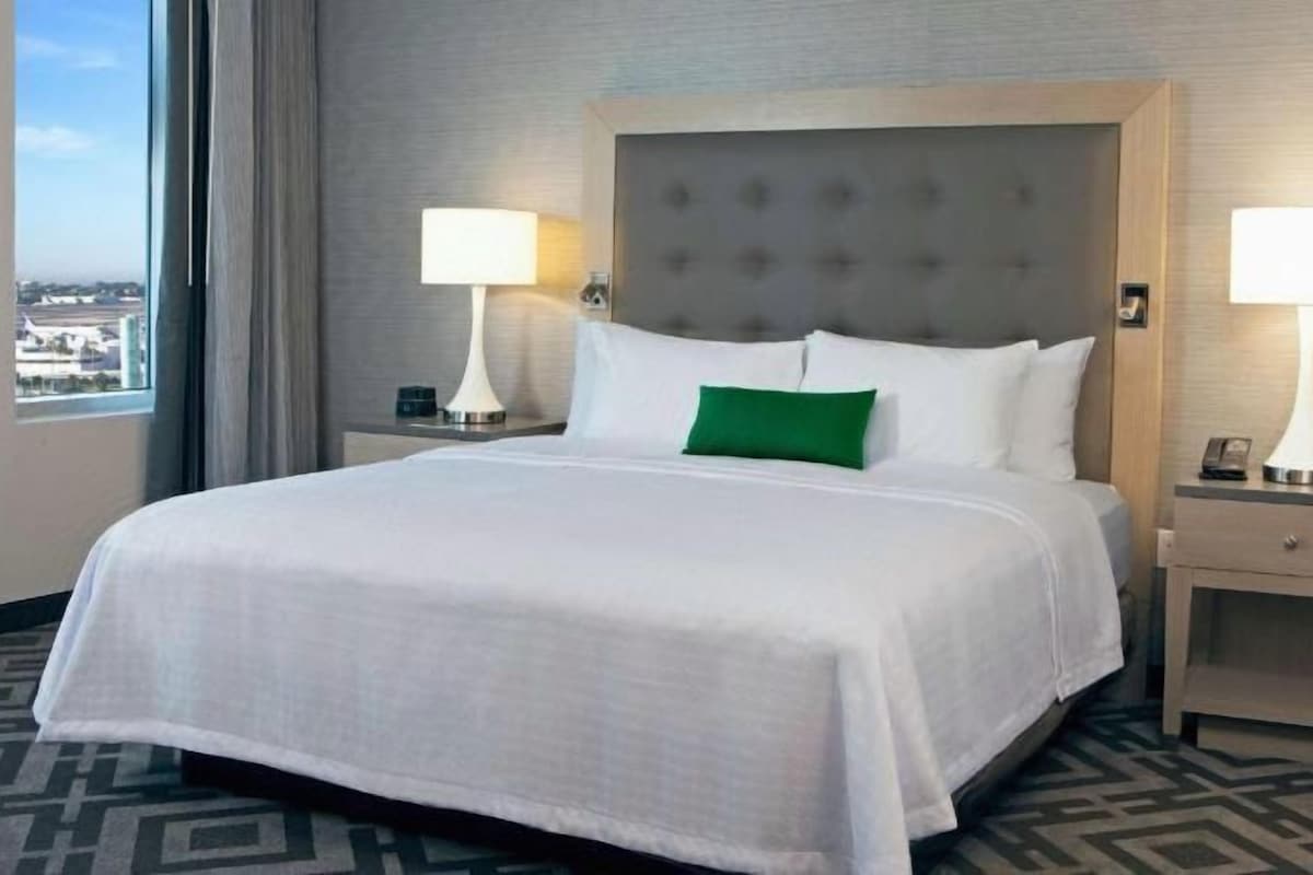A hotel room with a large bed, two bedside tables with lamps, a green pillow, and a view of the city.