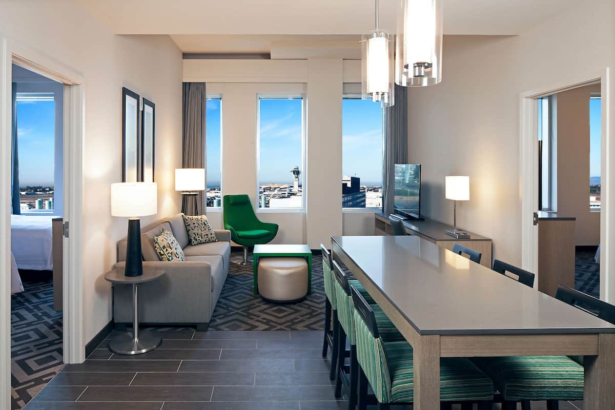 Ein modernes Hotelzimmer mit Essbereich, einer Couch, einem grünen Sessel und einem großen Fenster mit Blick auf die Stadt.