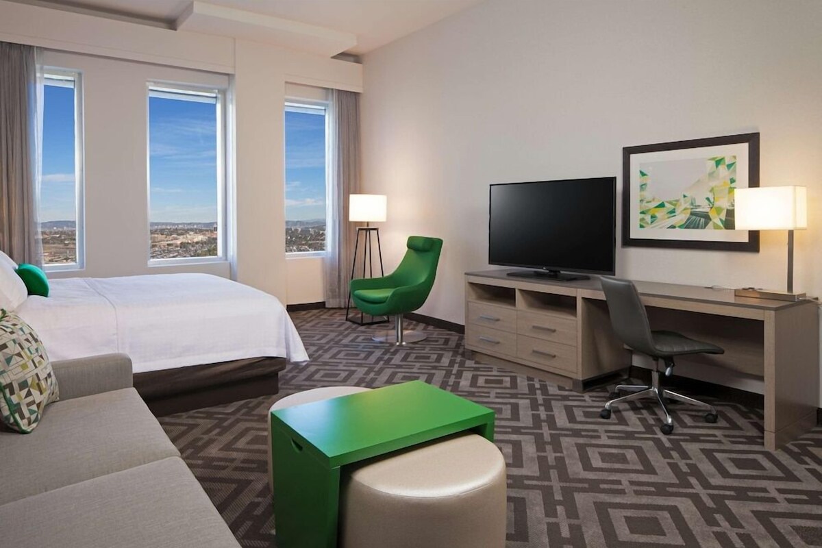 A hotel room with a bed, a desk, a television, a chair, and a view of the city.
