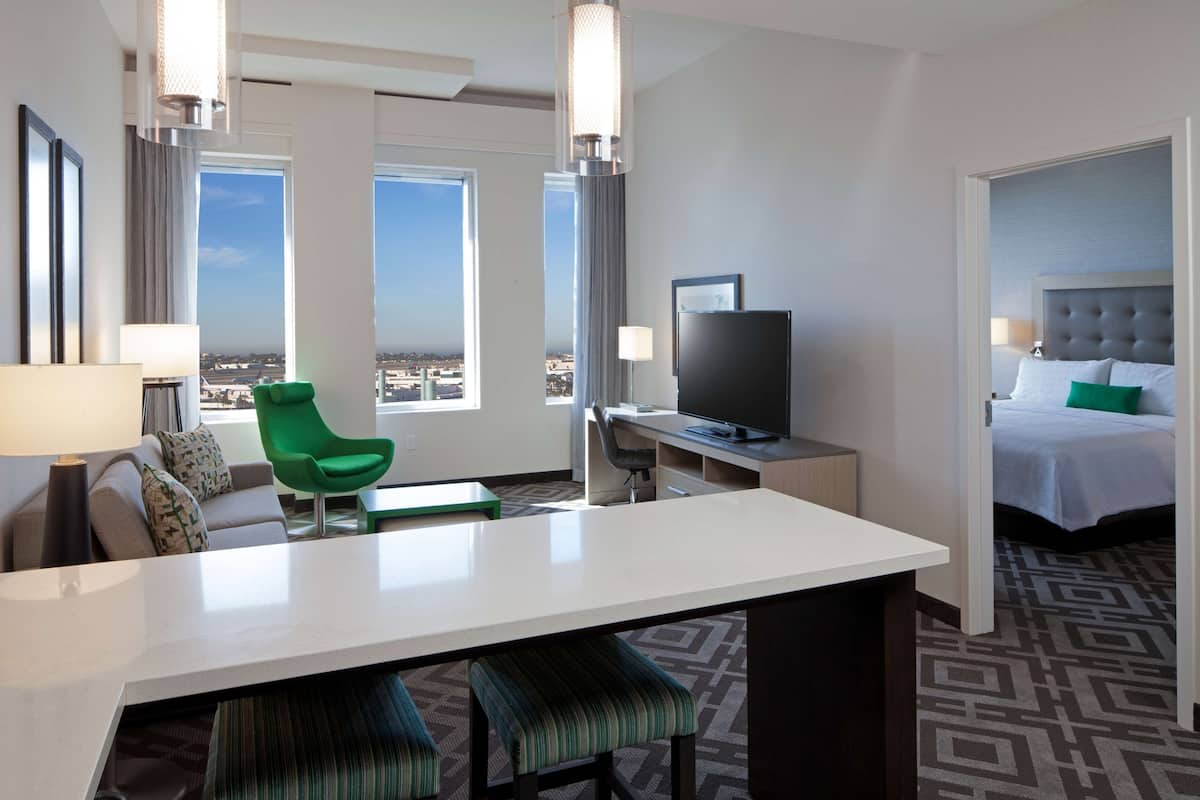 Ein Hotelzimmer mit einem großen Bett, einem Schreibtisch mit Fernseher, einem grünen Sessel und Blick auf die Stadt.