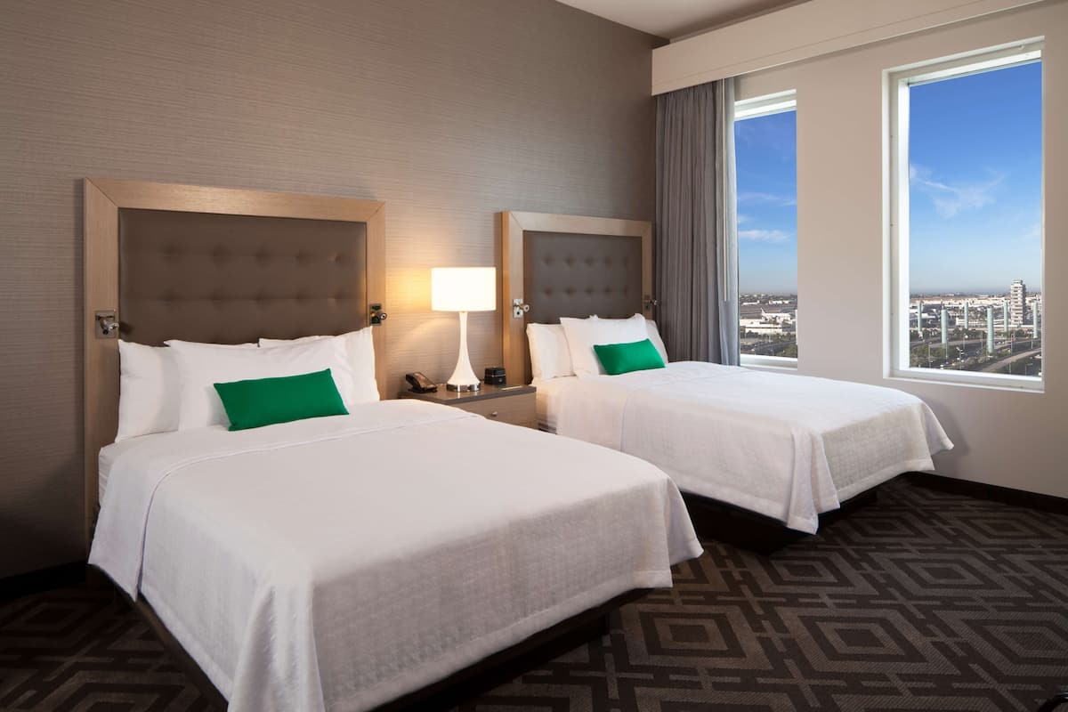Ein Hotelzimmer mit zwei Betten, einem großen Fenster und Blick auf die Stadt sowie einem gemusterten Teppich.