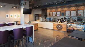 Una cafetería moderna con un mostrador central, zona de asientos con sillas moradas y carteleras en la pared.