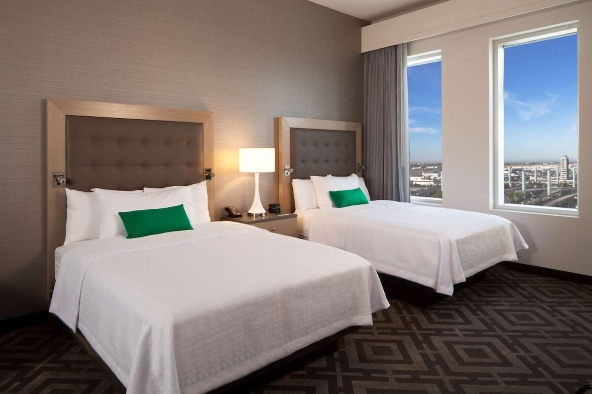 A hotel room with two beds, a large window with a city view, and a patterned carpet.