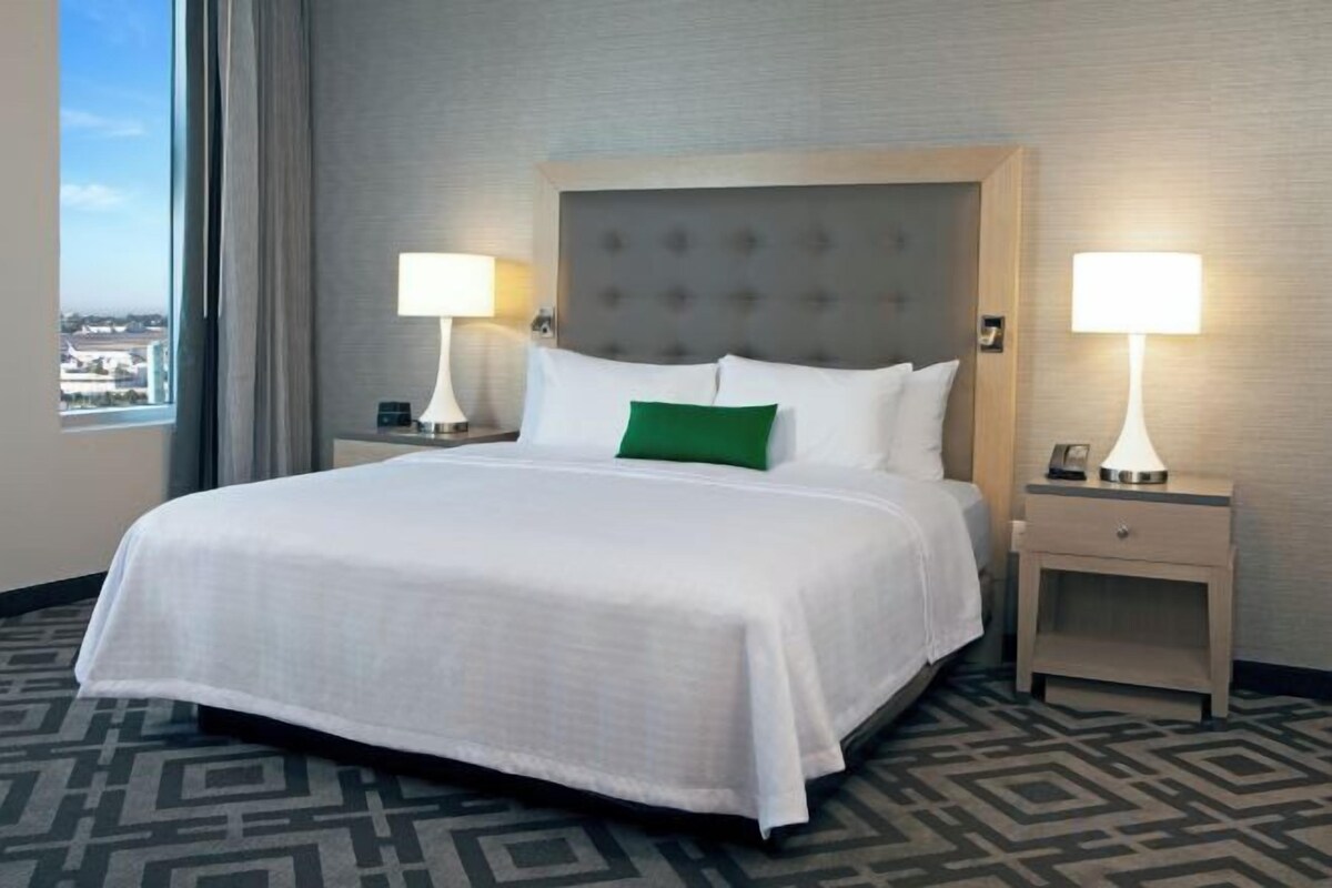 A hotel room with a large bed, two bedside tables with lamps, a view of the city, and a patterned carpet.