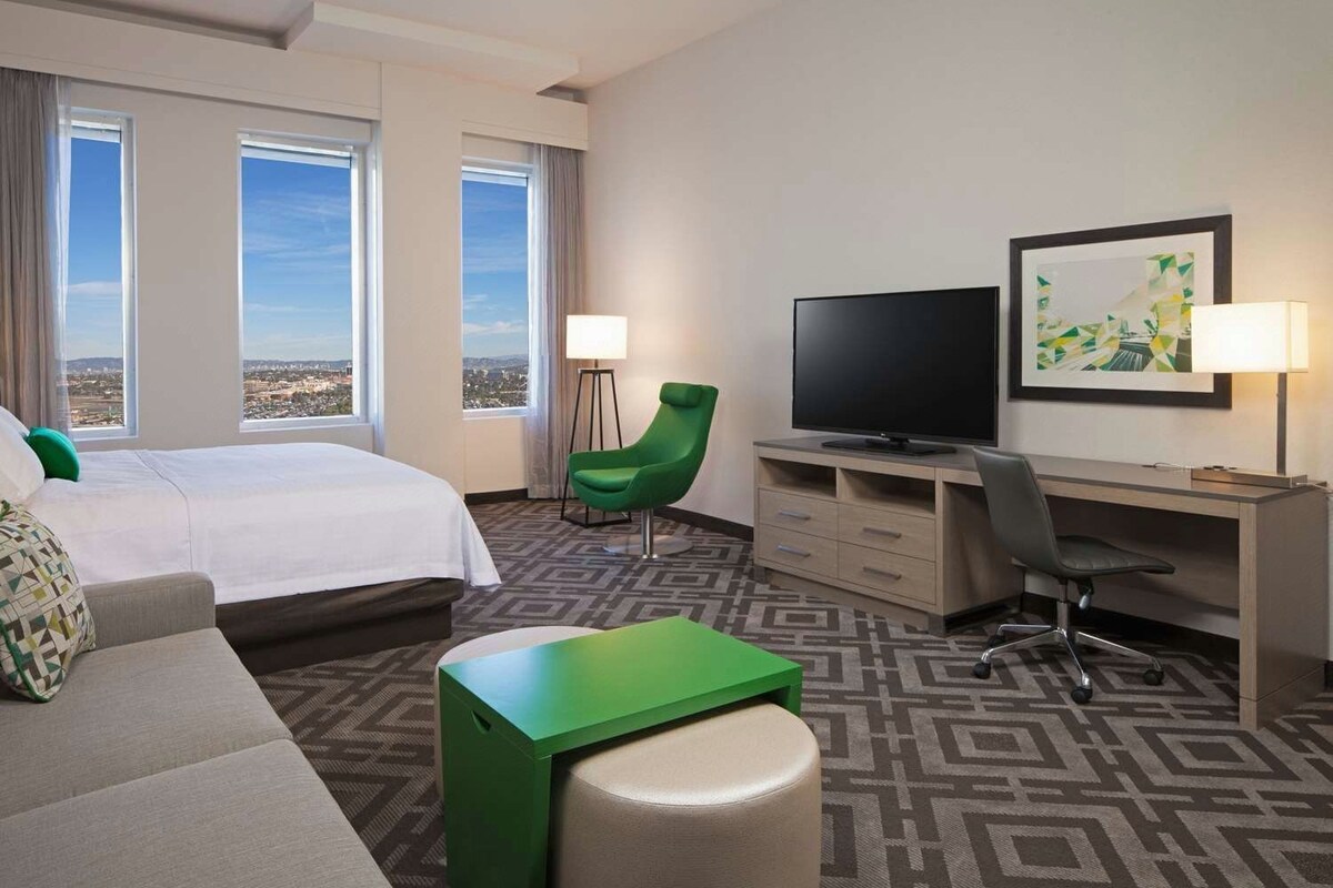 A hotel room with a bed, a desk, a television, a chair, and a view of the city.