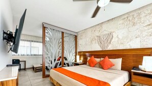 Une chambre d’hôtel avec un lit, une tête de lit en bois, des oreillers orange, un téléviseur fixé au mur et un ventilateur de plafond.
