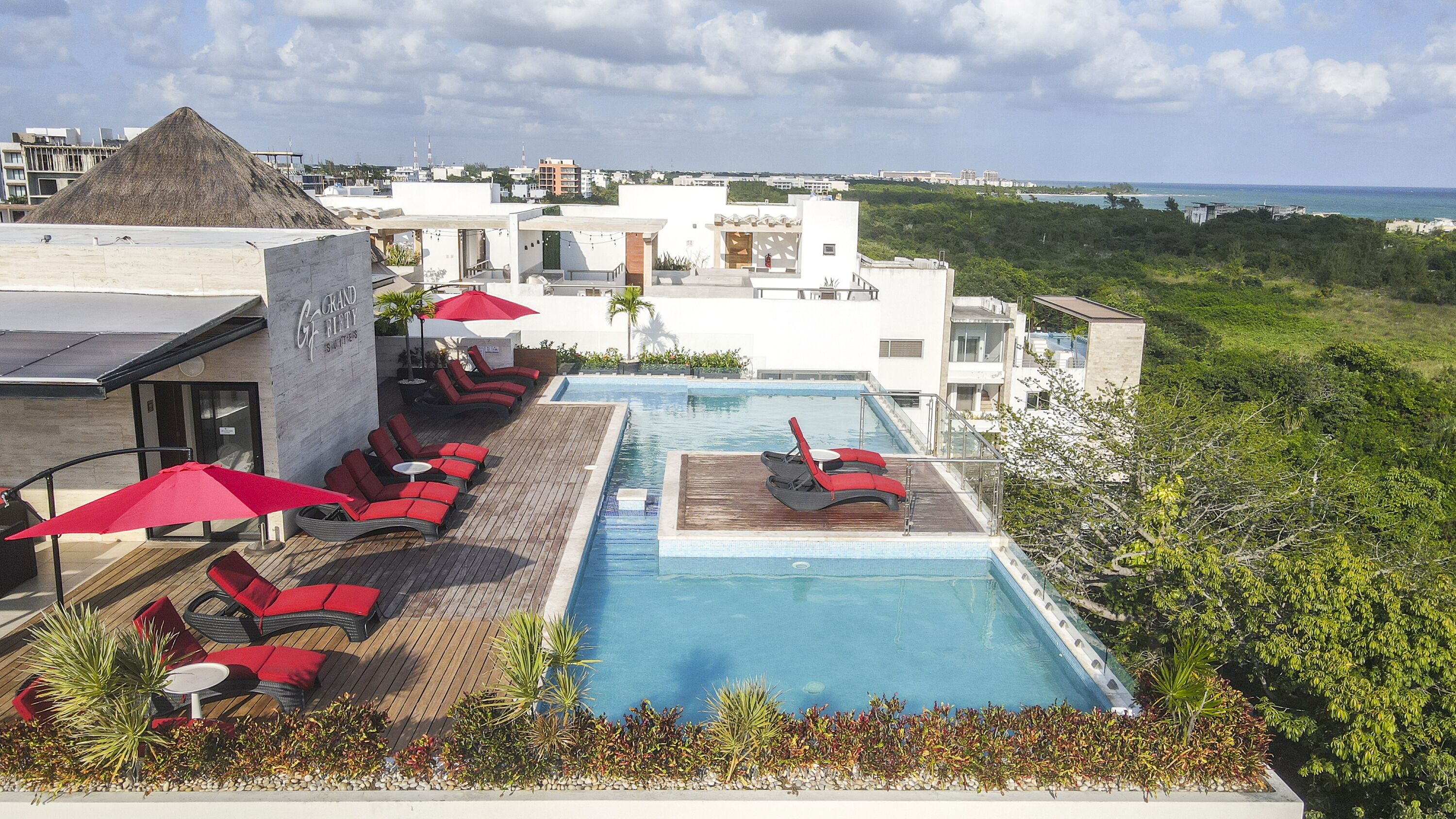 Un espace piscine aménagé sur le toit, avec des chaises longues rouges et des parasols, entouré de verdure et offrant une vue sur le paysage urbain.