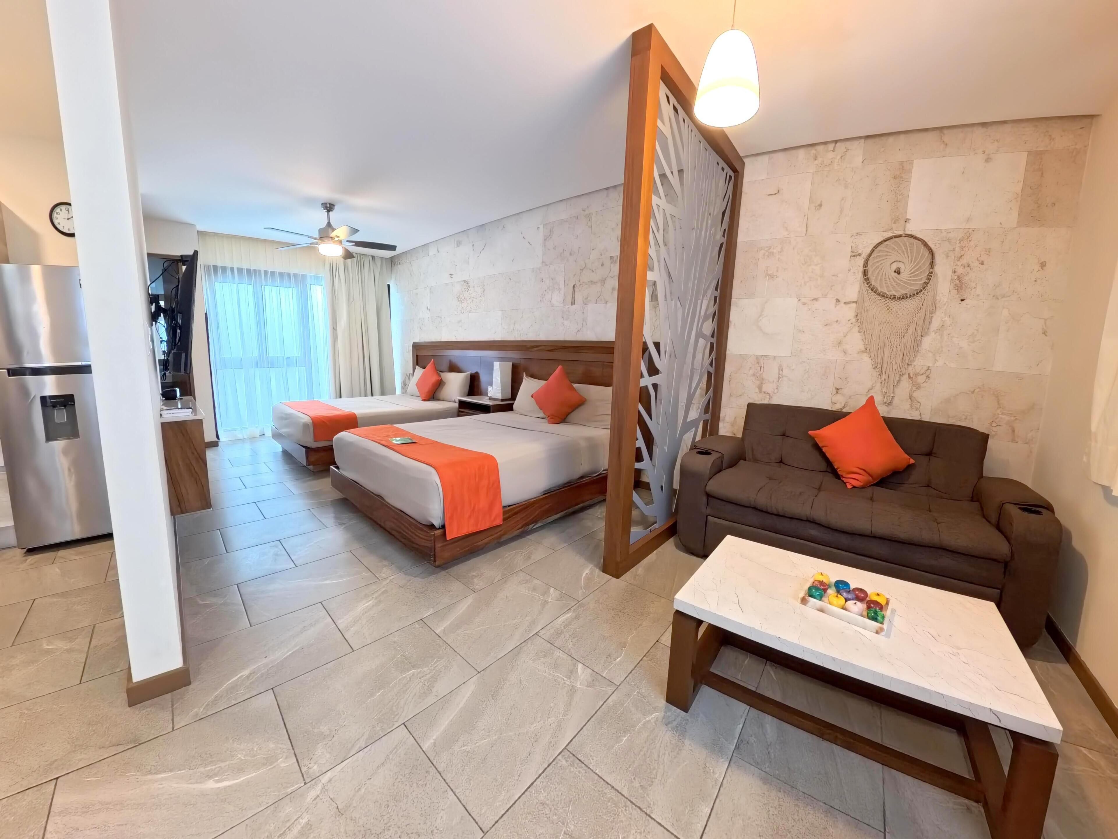 A hotel room with two beds, a sofa, a coffee table, a refrigerator, a ceiling fan, and a decorative wall piece.