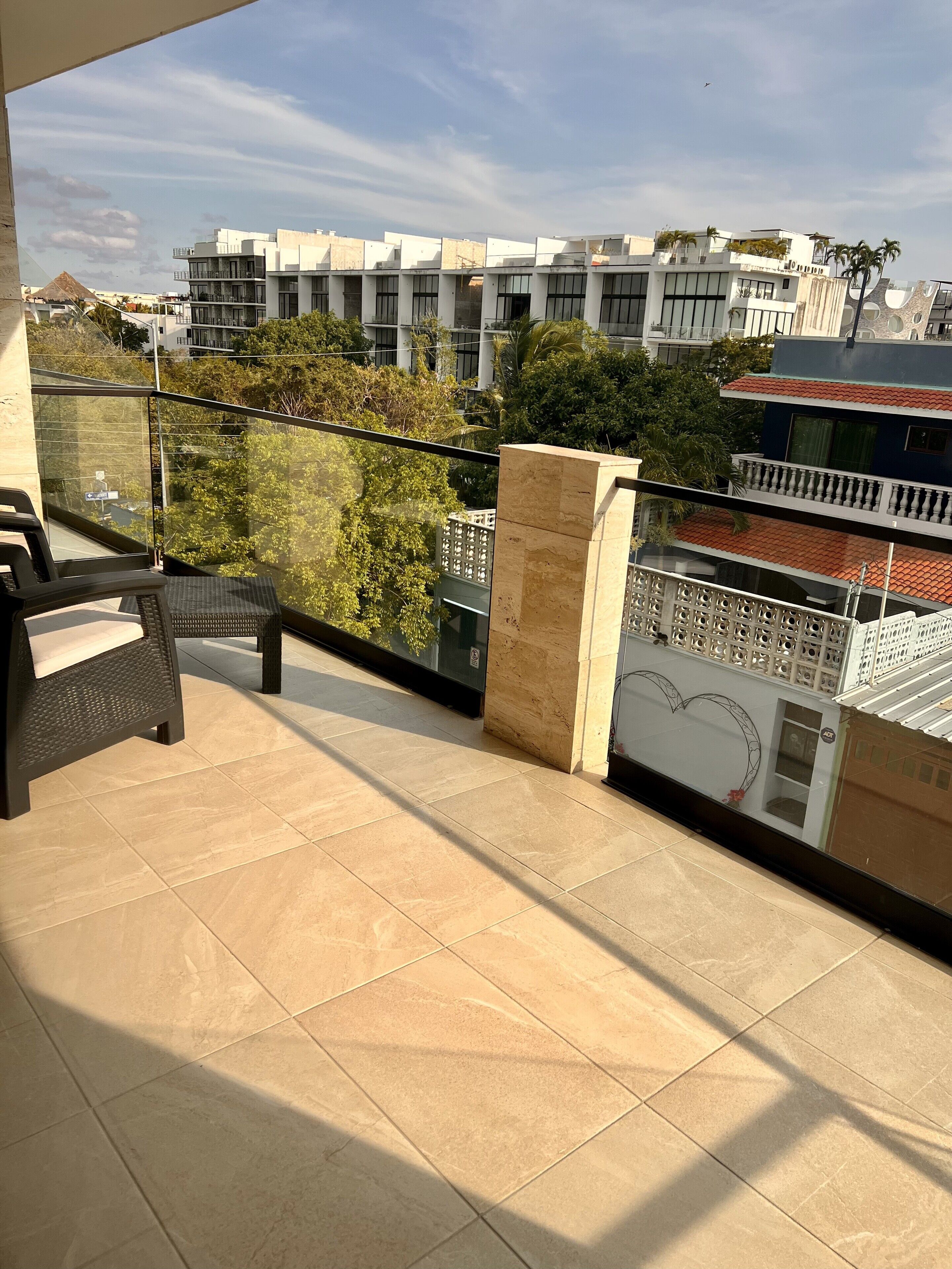 A balcony with tiled flooring, a glass railing, and a view of modern buildings and greenery.