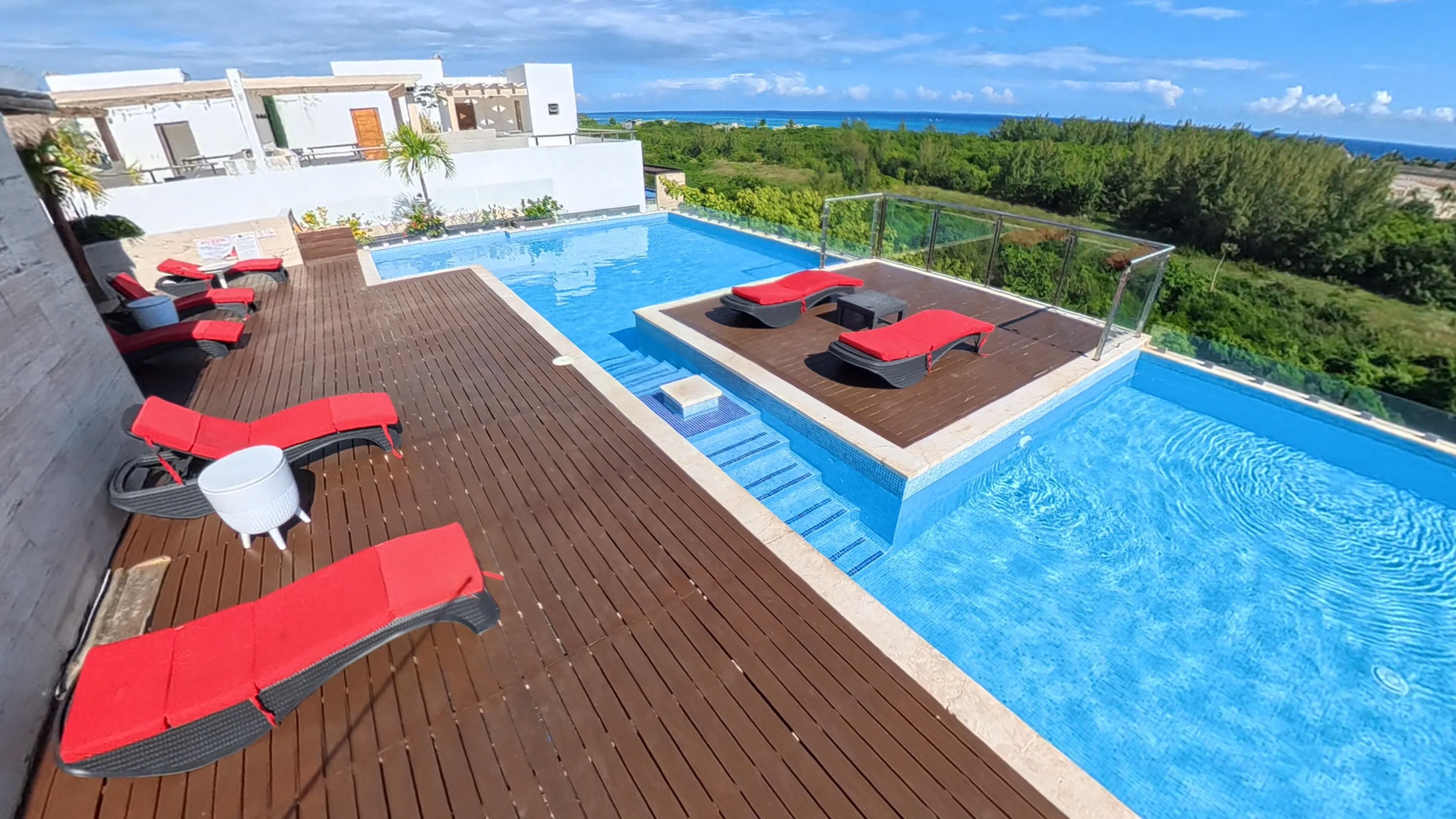 A rooftop pool with red loungers, a wooden deck, and a view of the ocean.