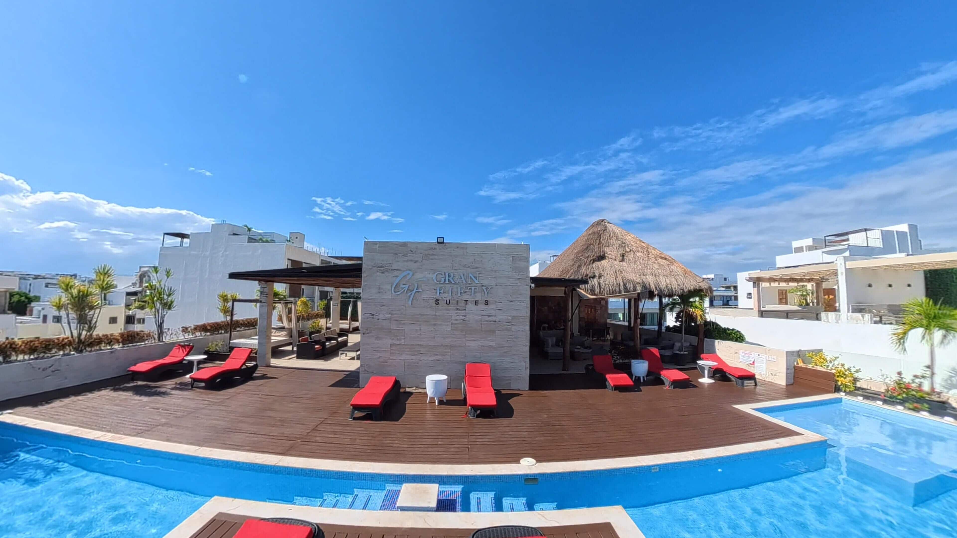 Un área de piscina en la azotea con sillones de descanso, una estructura con techo de paja y un letrero que dice 'GRAND FIFTY SUITES'.
