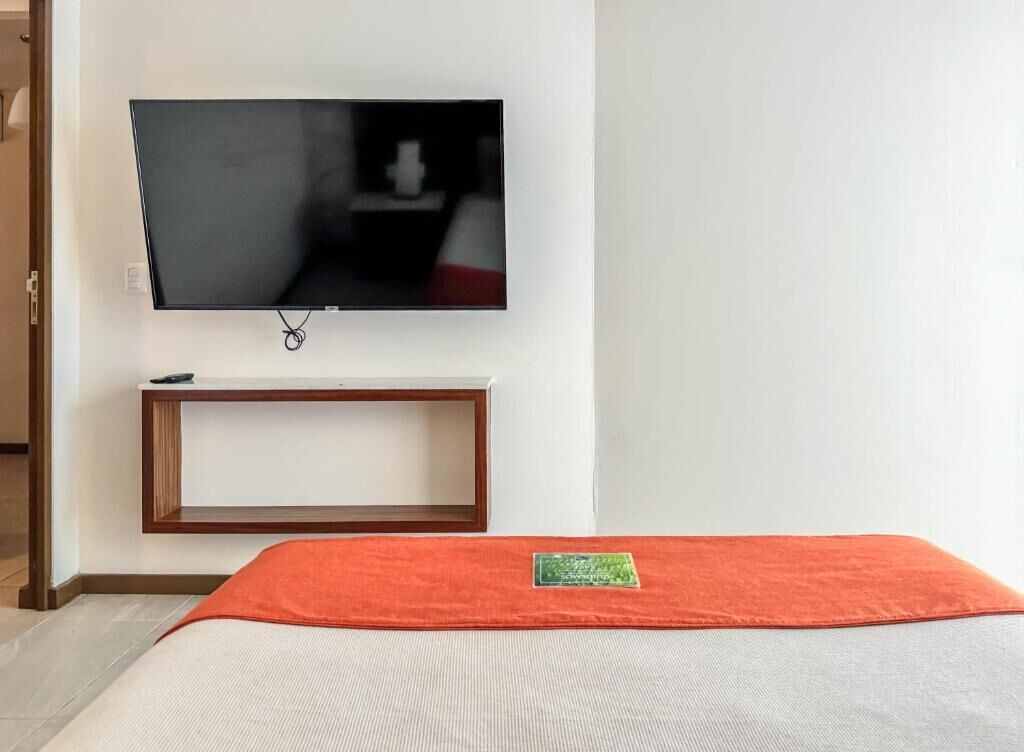 A room with a bed, a television mounted on the wall, and a small wooden shelf below it.