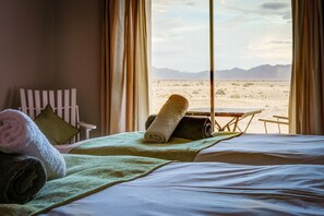 A hotel room with a bed, a chair, and a view of a desert landscape through large windows. - Elegant Desert Lodge (Sesriem)