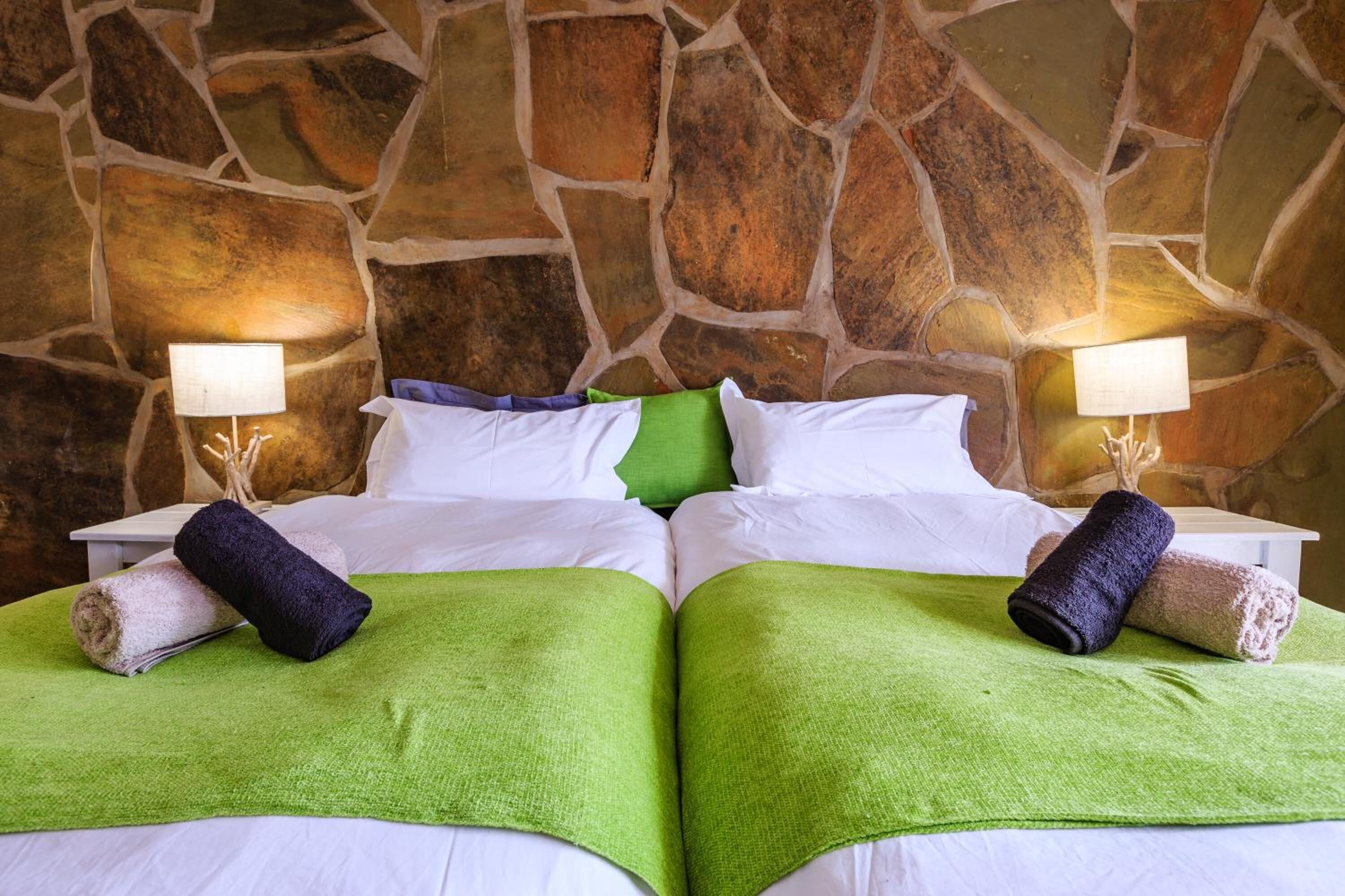 A double bed with green and white linens, two rolled towels, and a stone wall backdrop.