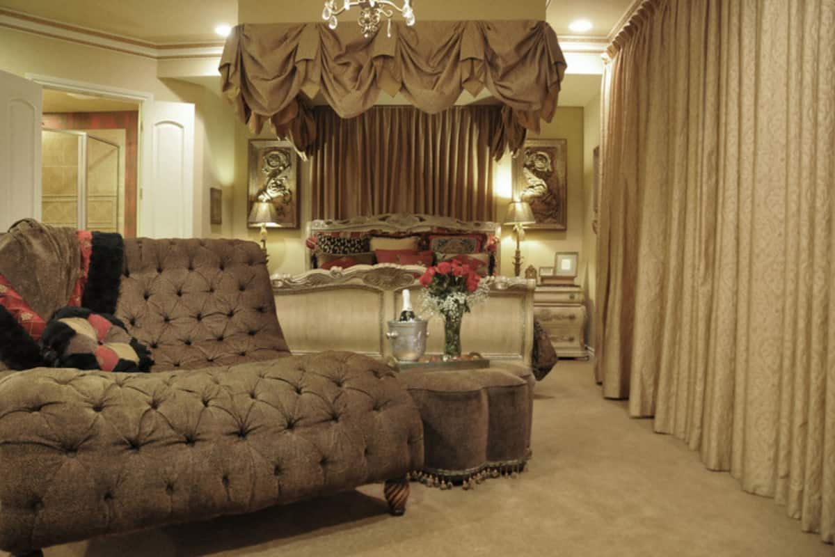 A luxurious bedroom with a tufted sofa, a large bed with ornate bedding, a nightstand with a lamp, and a vase of flowers.