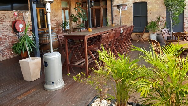An outdoor dining area with a long wooden table and chairs, surrounded by potted plants and a metal heater.