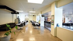 A spacious cafeteria with long tables and chairs, a large potted plant, and a counter with various items.