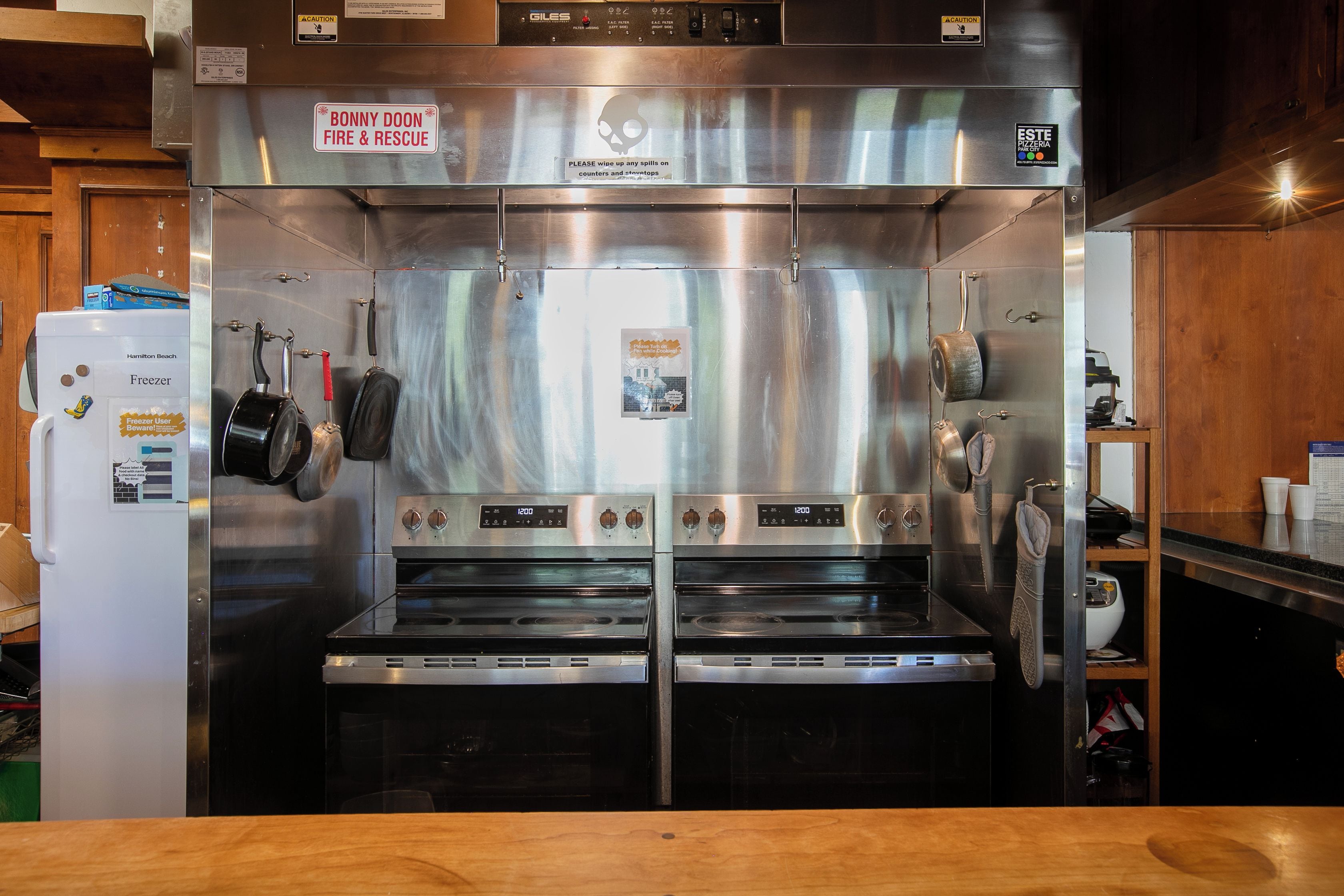 A commercial kitchen with stainless steel appliances, including a freezer, two ovens, and a warning sign for Bonny Doon Fire & Rescue.