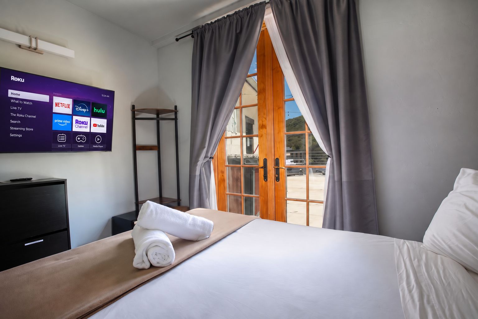 A hotel room with a bed, a TV displaying Roku interface, a wooden ladder, and a view through a glass door.
