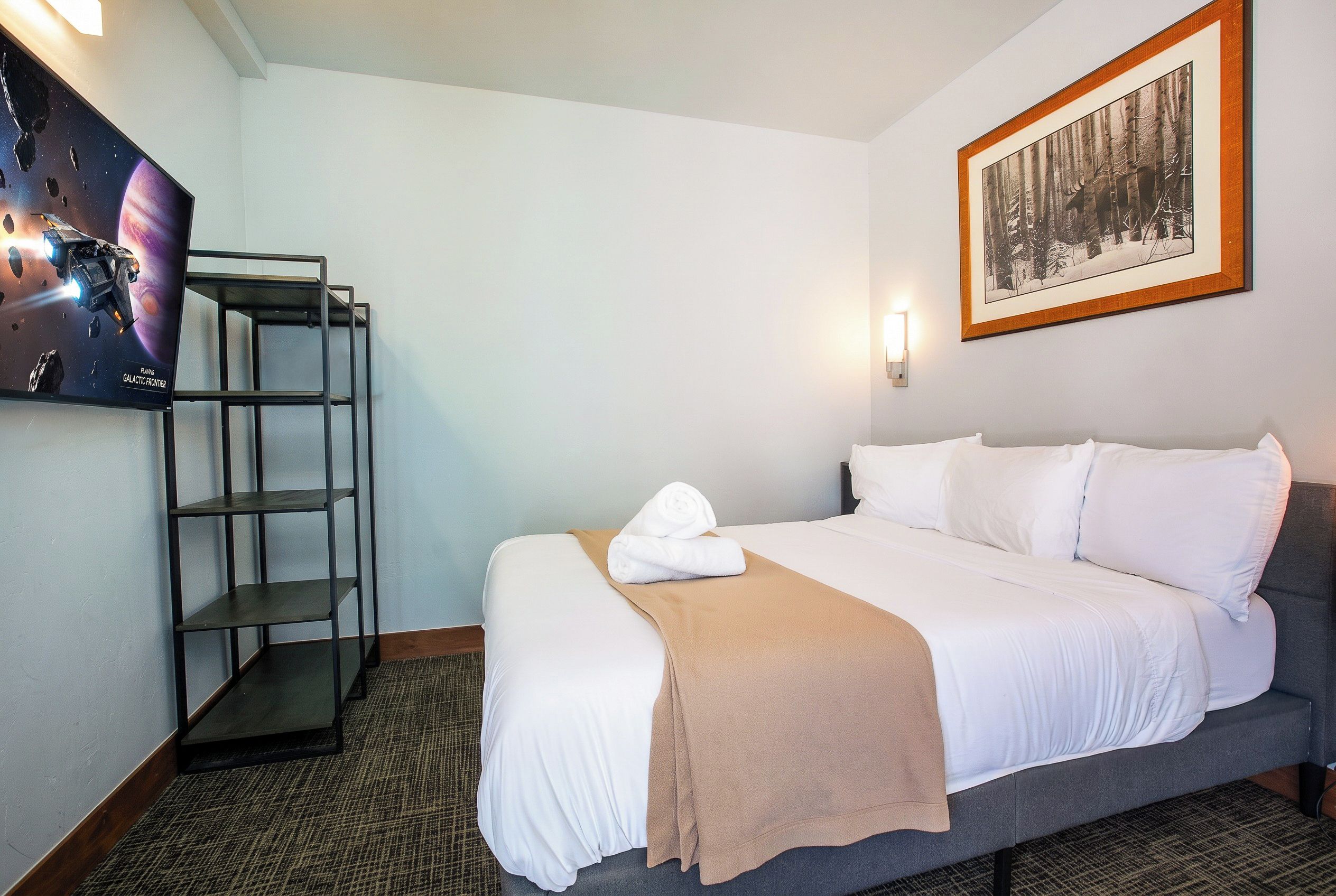 A hotel room with a bed, a television mounted on the wall, a framed picture, and a shelf unit.