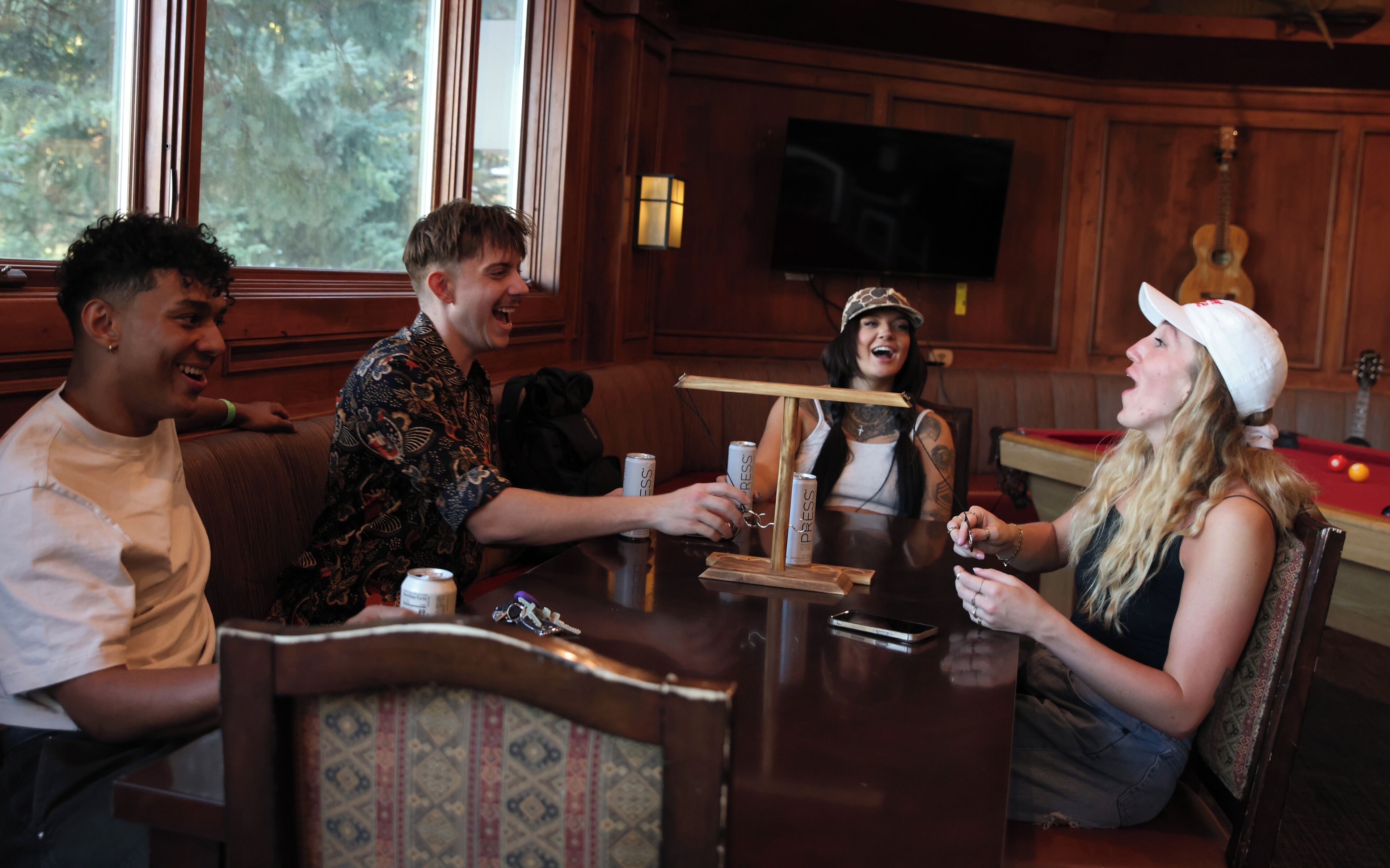 A group of people sitting around a wooden table in a room with a pool table and a television mounted on the wall.