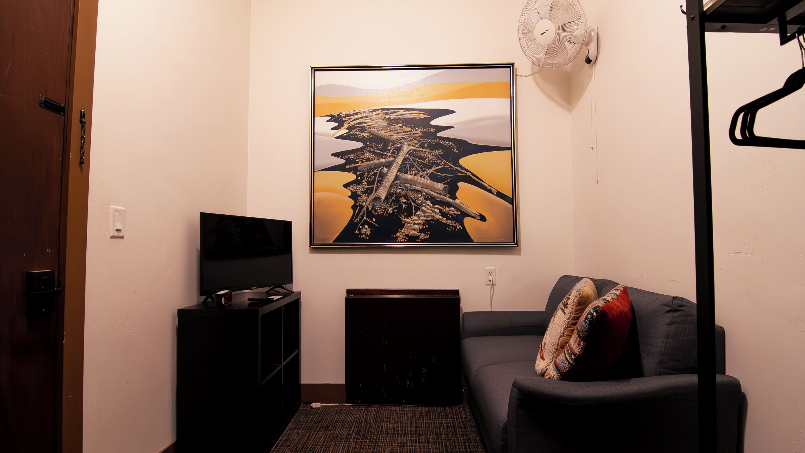 A room with a flat-screen TV, a framed abstract painting, a small speaker, a sofa with a patterned cushion, and a wall-mounted fan.