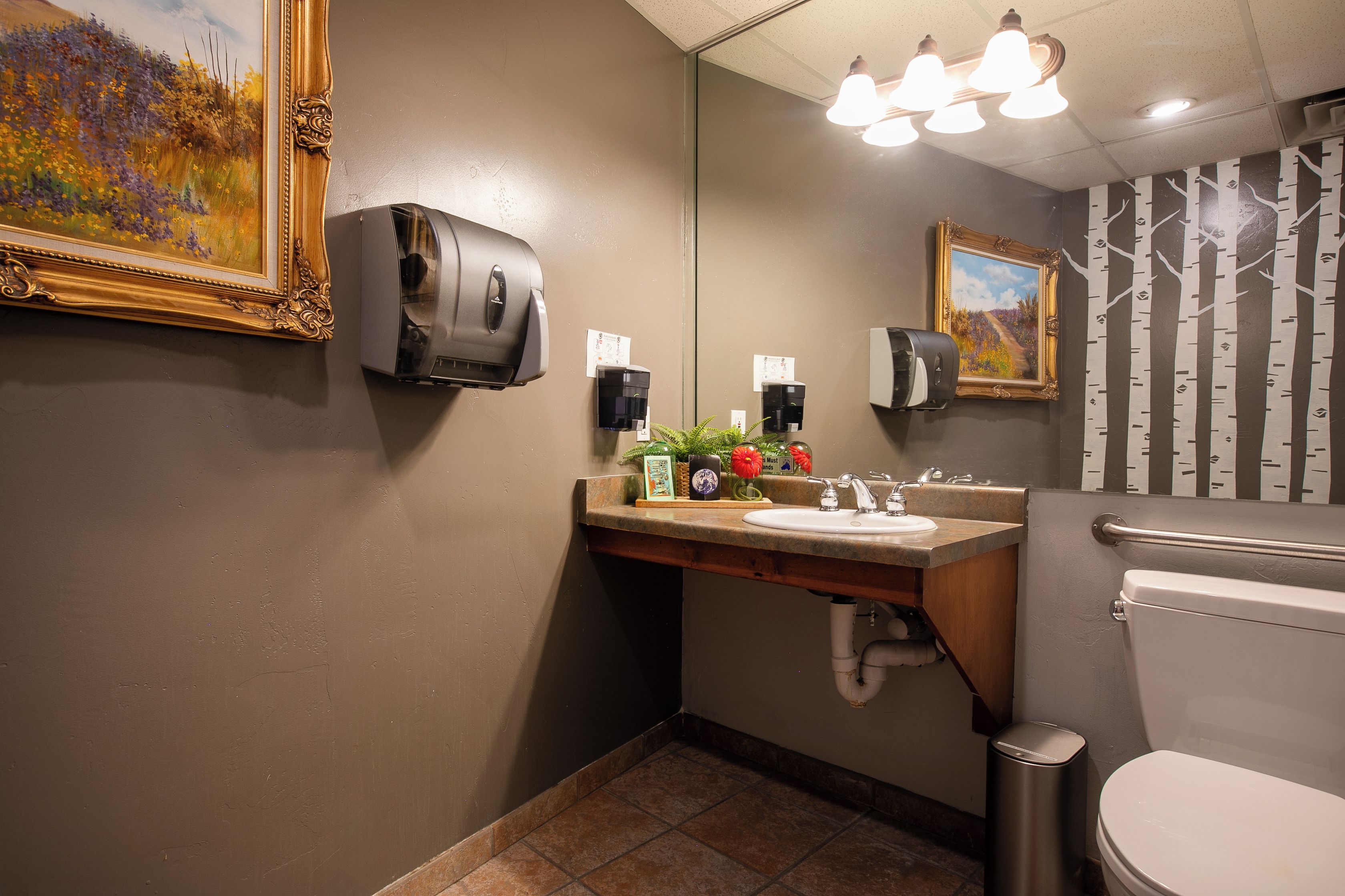 Un bagno con lavandino, specchio, asciugatore ad aria e WC. Sono presenti erogatori a parete e un quadro incorniciato sopra lo specchio.
