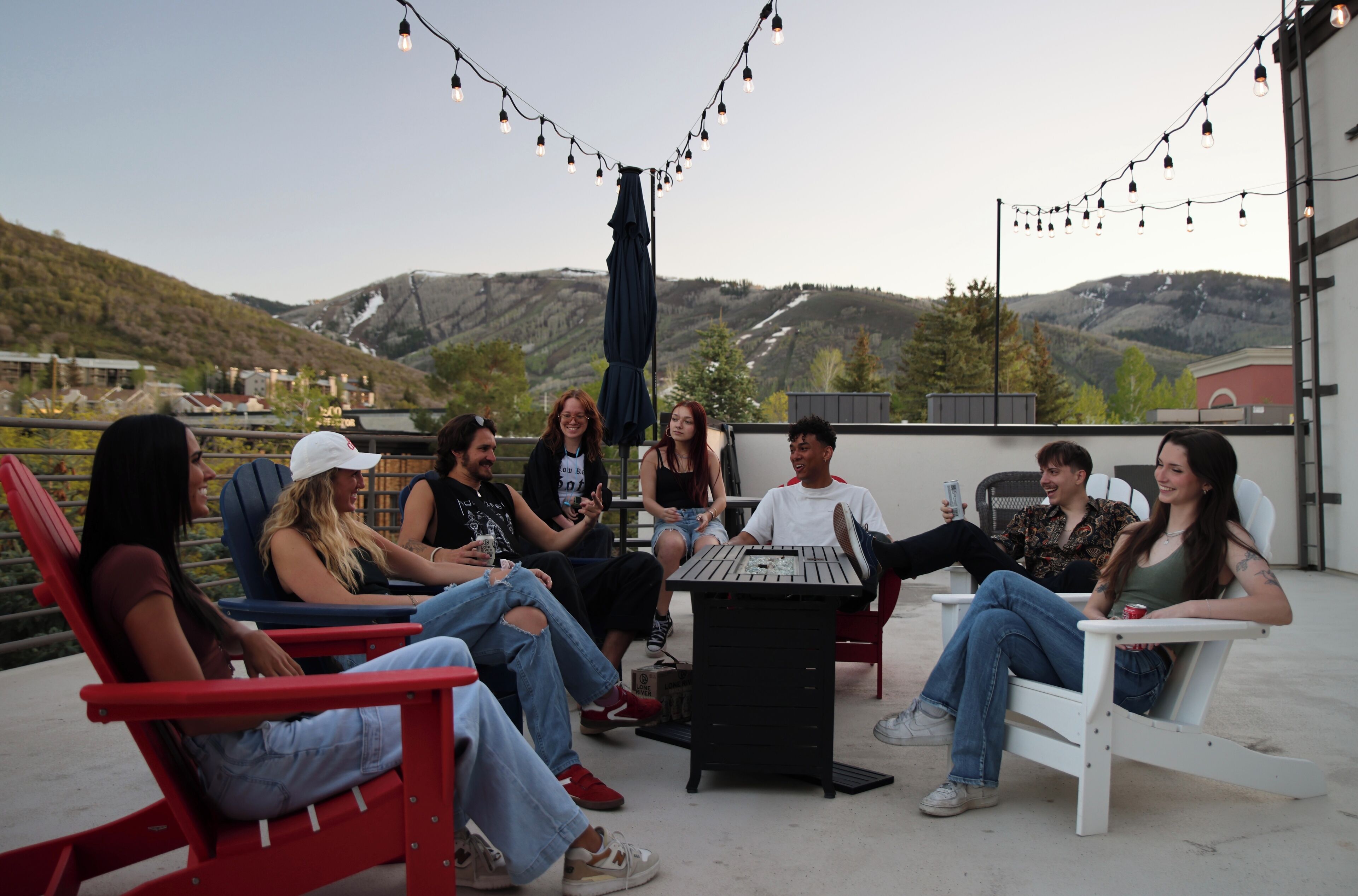 A group of people seated on outdoor chairs and sofas around a table on a rooftop terrace with string lights overhead.