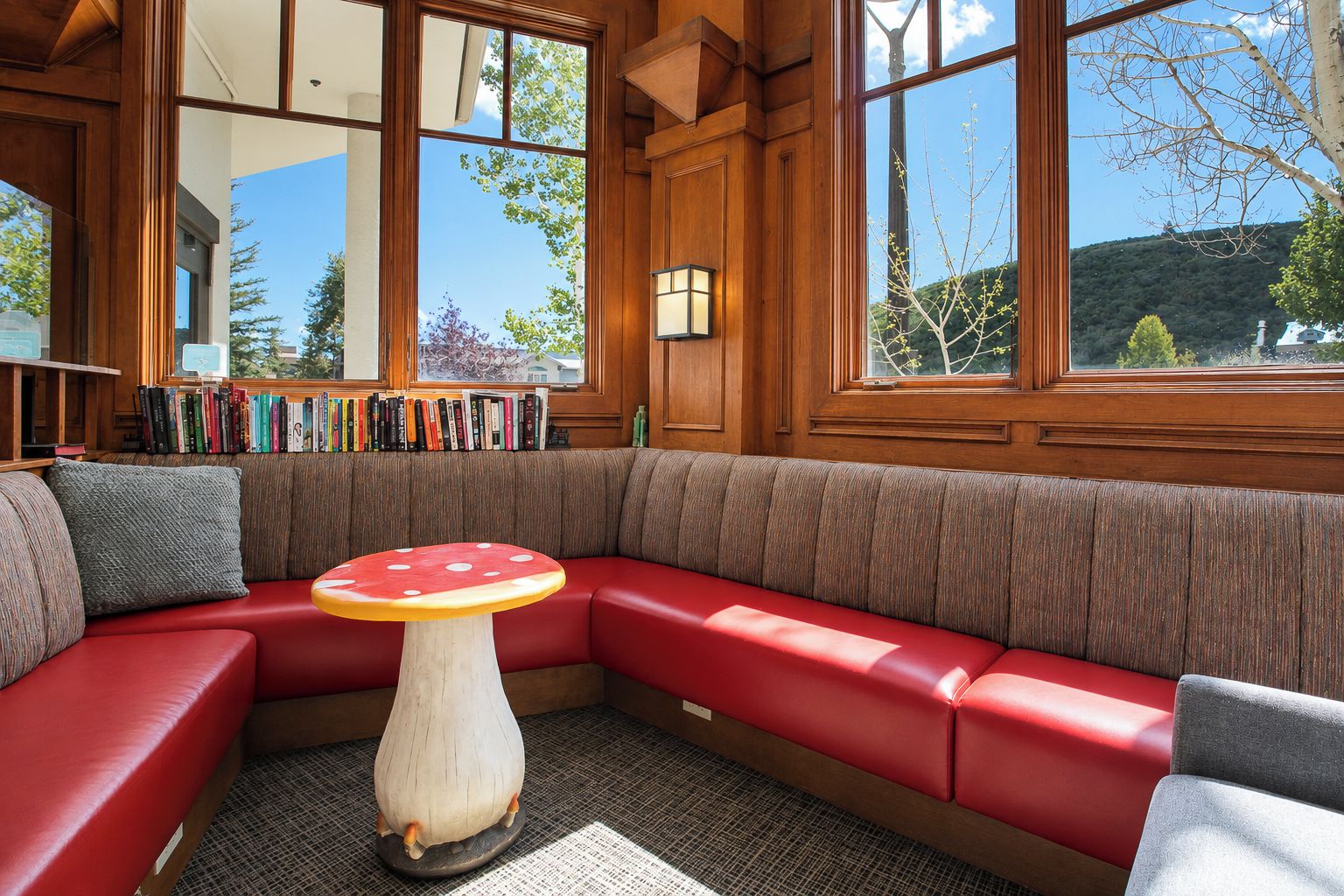 Un coin lecture confortable avec un banc rembourré de couleur rouge, une table en bois ornée d’un motif en forme de champignon et une bibliothèque remplie de livres.