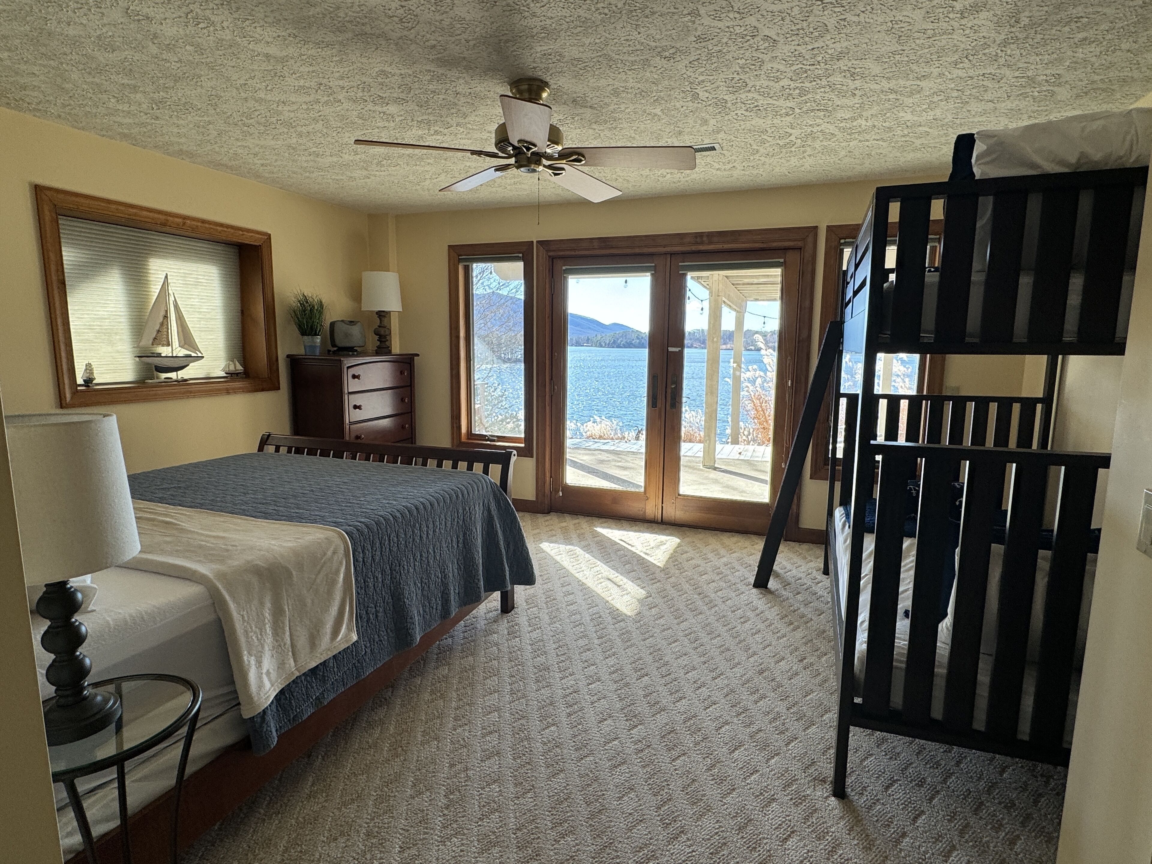 Une chambre à coucher avec un lit, une commode, un ventilateur de plafond et une vue sur un lac, visible à travers une porte vitrée.