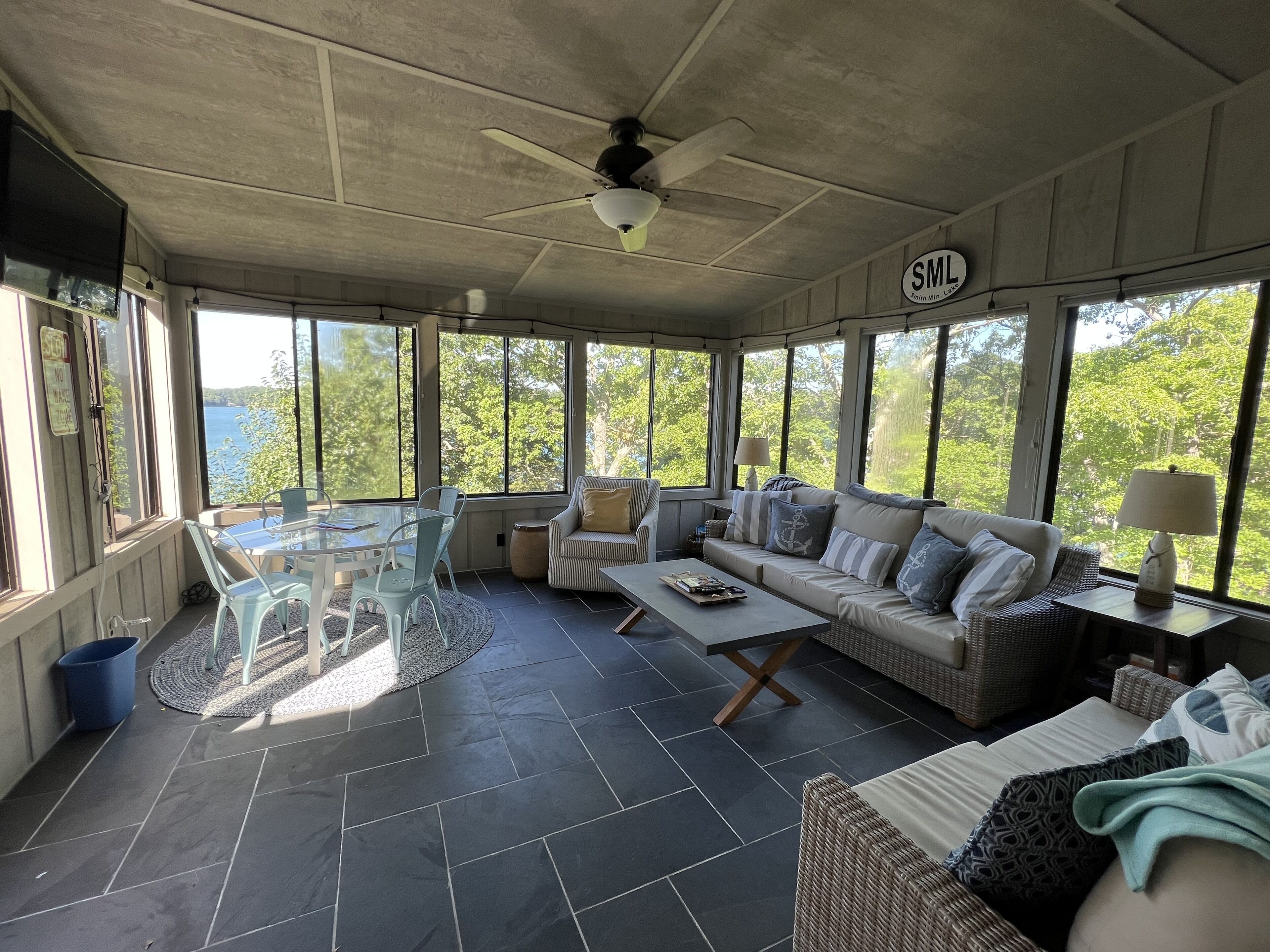 Une pièce lumineuse, baignée de soleil, avec une grande fenêtre, un ventilateur de plafond, un canapé, une table basse et un fauteuil en osier.