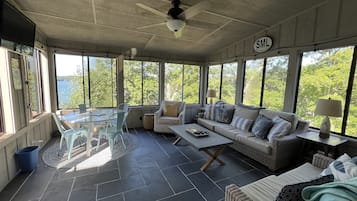 Une pièce lumineuse, baignée de soleil, avec une grande fenêtre, un ventilateur de plafond, un canapé, une table basse et un fauteuil en osier.