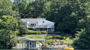 Una casa frente al lago con muelle, un bote cubierto y una pequeña terraza con una estatua.