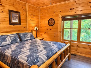 A wooden cabin room with a bed, a nightstand, a lamp, a framed picture, and a window with a view of greenery. - Elevator + Roll-In Shower | Theater + Game Room (Pigeon Forge)