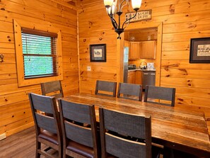 A wooden interior with a dining table and chairs, a kitchen area, and a window with blinds.