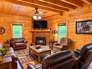 A cozy log cabin interior with a fireplace, leather furniture, a television, and a bear painting. - Elevator + Roll-In Shower | Theater + Game Room (Pigeon Forge)