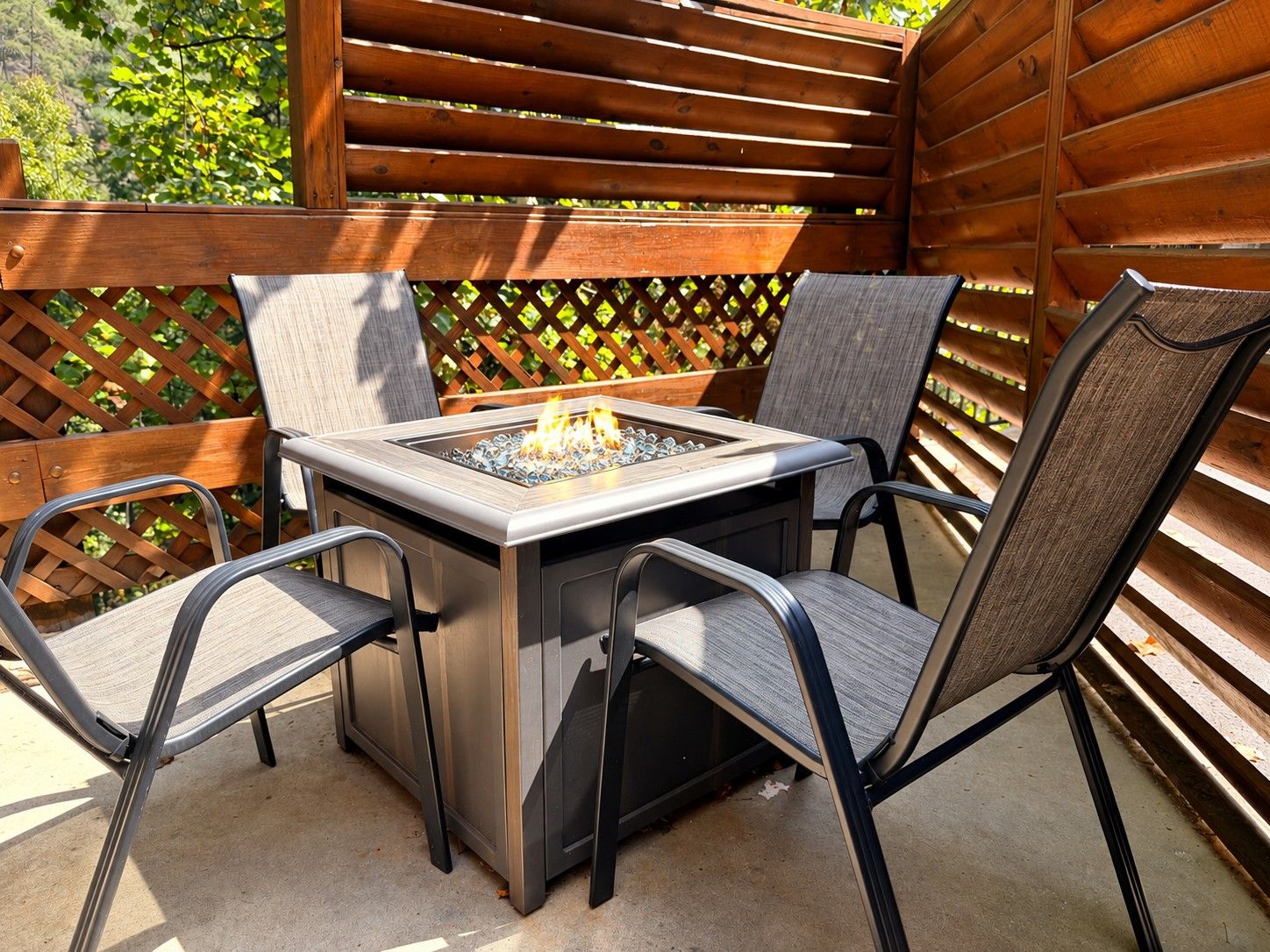 An outdoor patio with a square table and four chairs, a fire feature in the center, and a wooden lattice backdrop.