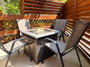 An outdoor patio with a square table and four chairs, a fire feature in the center, and a wooden lattice backdrop.