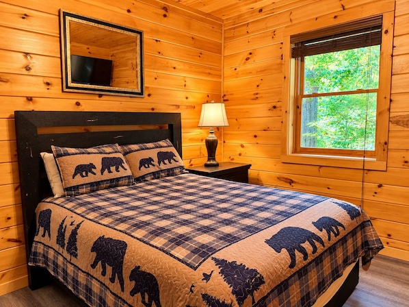 A wooden cabin bedroom with a bed, a nightstand, a lamp, a mirror, and a window.