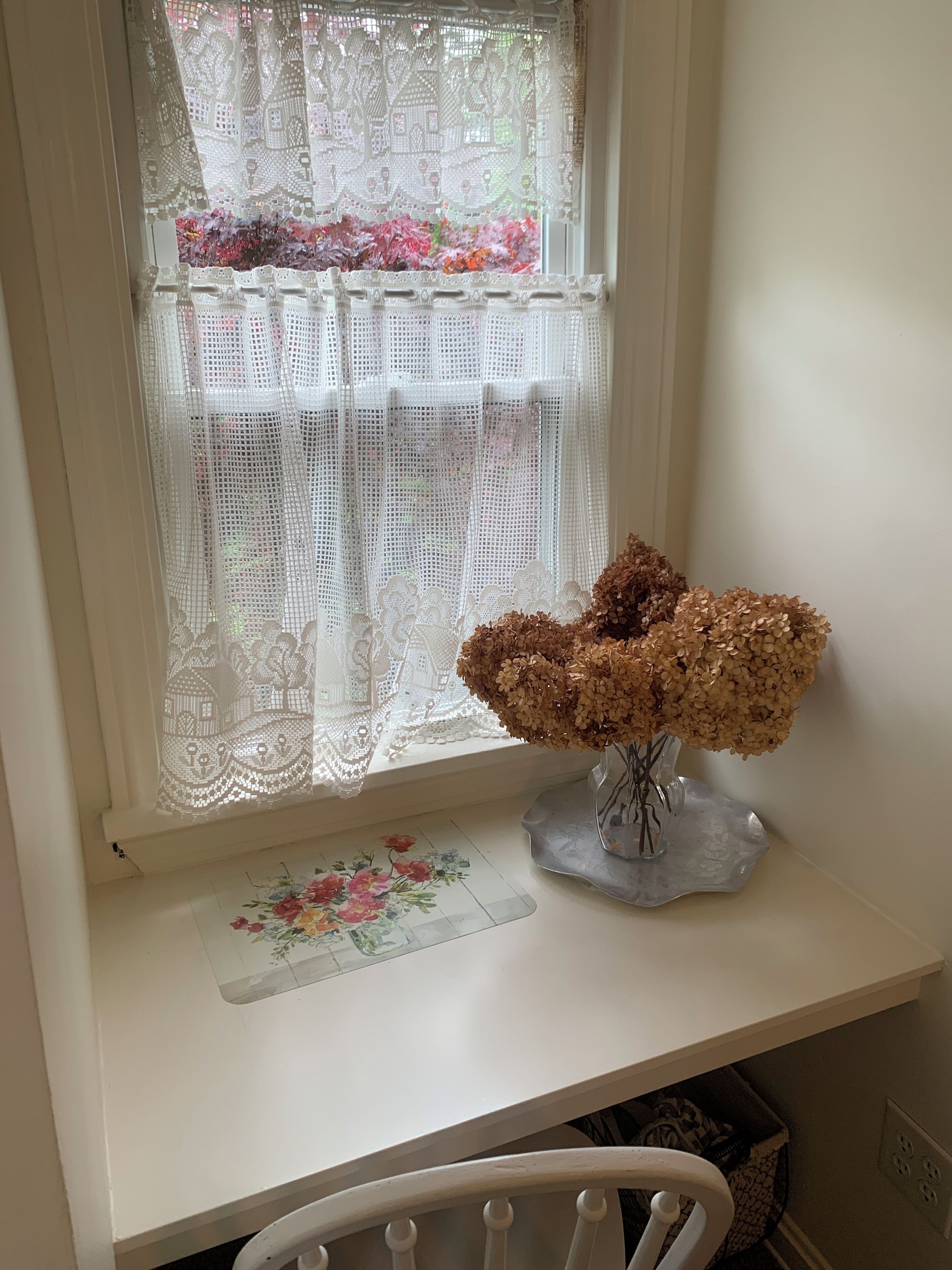 A window with lace curtains, a vase with dried flowers, and a floral mat on a white shelf.