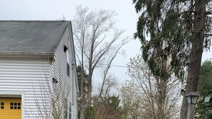A house with a yellow garage door, a brick pathway leading to the entrance, and a garden with green plants and a tree. - Cozy Country Apartment in Heart of Geauga County (Newbury)