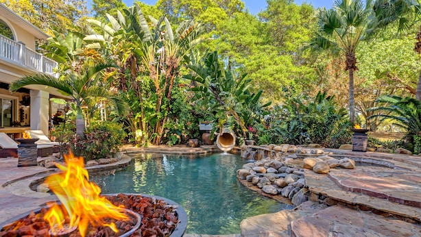 Zona de piscina con una chimenea escultural, rodeada de plantas tropicales y un suelo de piedra.