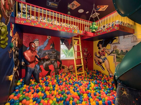 A playroom with a ball pit, superhero murals, and a slide.