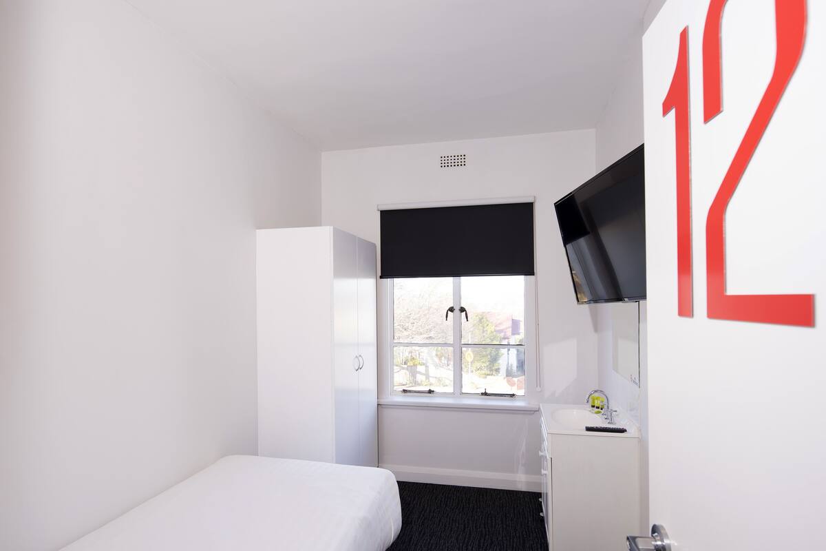 Une petite chambre moderne avec un lit simple, une fenêtre munie d’un store noir, une armoire blanche, un petit bureau avec un miroir et un téléviseur fixé au mur.