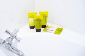 Une salle de bain avec un lavabo blanc, trois tubes jaunes étiquetés « ÉVOLUTION » pour le shampoing, le gel douche et le revitalisant, et un savon.