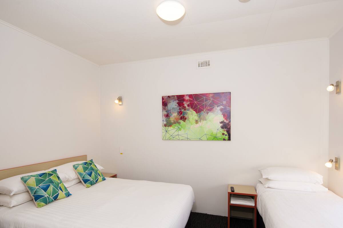 Une chambre d’hôtel avec un lit, deux oreillers, une table de chevet, un tableau accroché au mur et un luminaire au plafond.